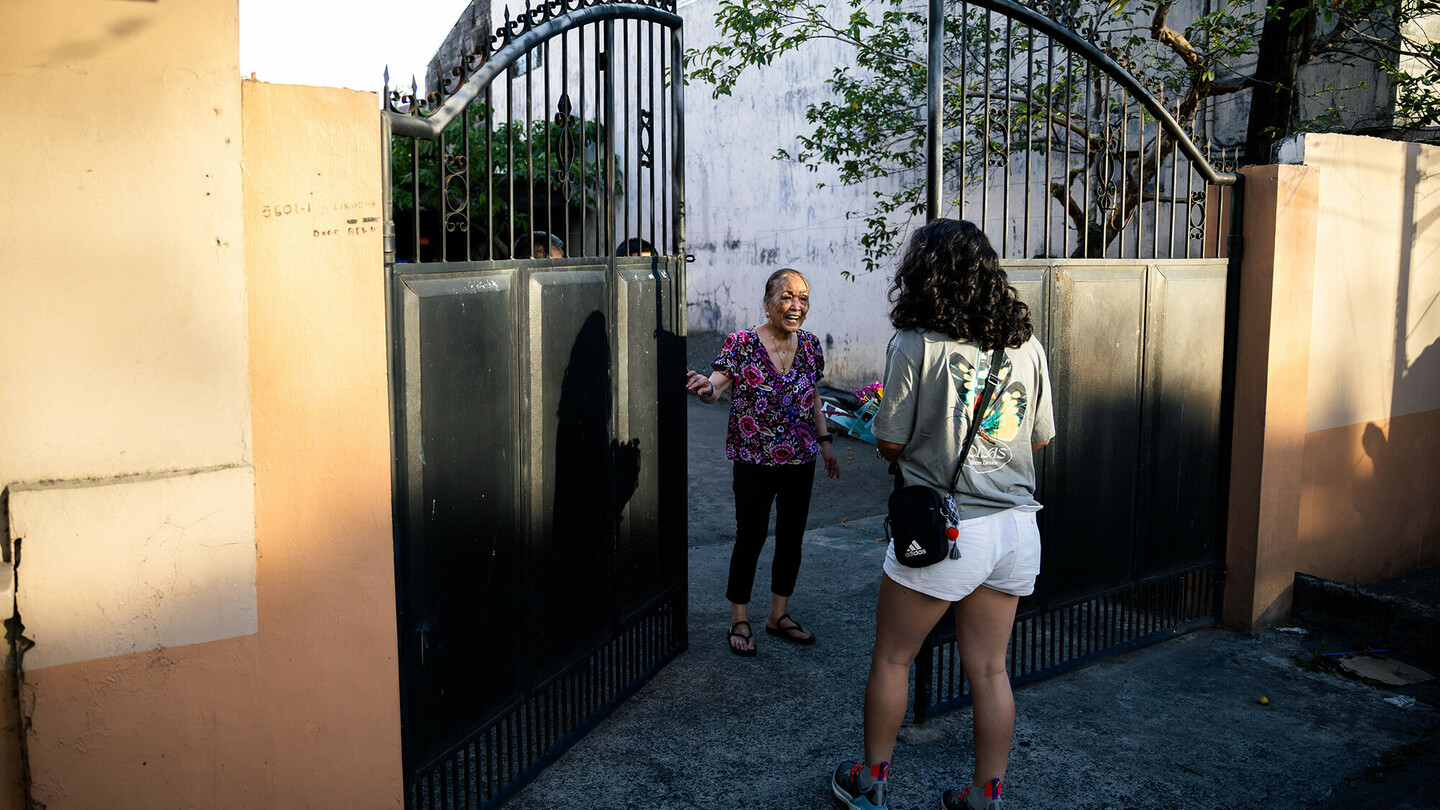 An older woman in a patterned top stands smiling at an open gate, talking to a younger woman who is looking at her. The scene takes place in an alley with house walls and a tree in the background.