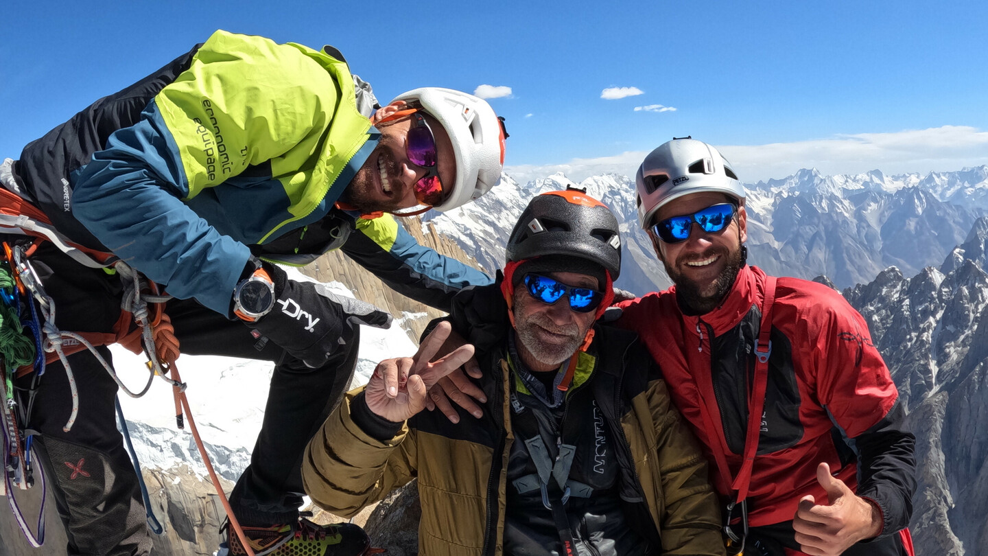 Three climbers, including Eduard Marín, on a snowy summit with climbing gear and helmets, smiling in front of a stunning mountain landscape.