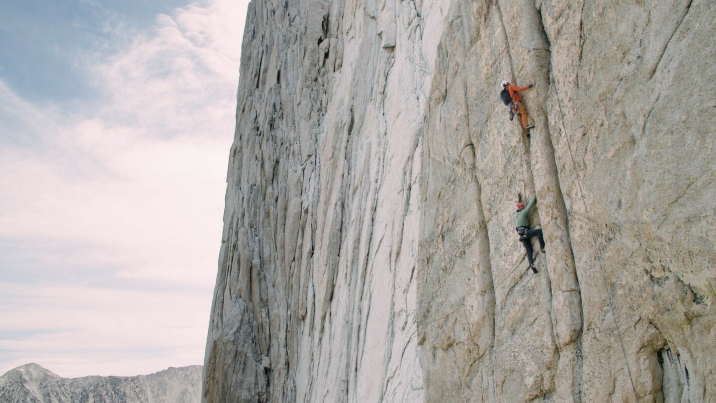 Two climbers in climbing gear and helmets are scaling a steep, vertical rock face. The upper climber is wearing an orange jacket, while the lower climber is in a green jacket. The massive wall rises high into the sky, set against a clear, slightly cloudy sky in the background.